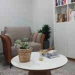 Detalhe da sala de atendimento, com poltrona, mesa de centro, estante de livros e objetos de decoração